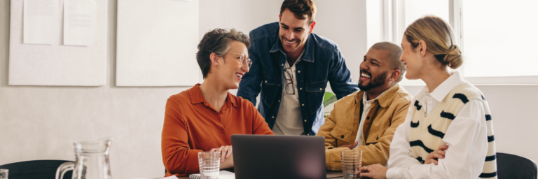 Four colleagues laugh and discuss around a laptop in a bright office setting, conveying a collaborative and cheerful work environment.