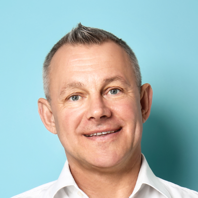 Middle-aged man with a mohawk hairstyle smiling against a turquoise background, wearing a white shirt.