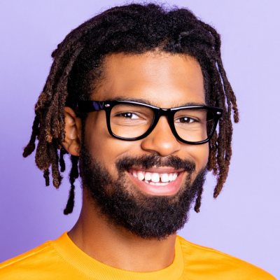 Portrait of a smiling man with glasses and dreadlocks, wearing a yellow shirt against a purple background.