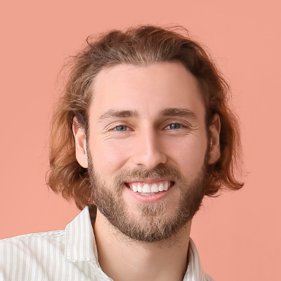 A smiling man with shoulder-length wavy brown hair and a beard, wearing a striped shirt against a pink background.