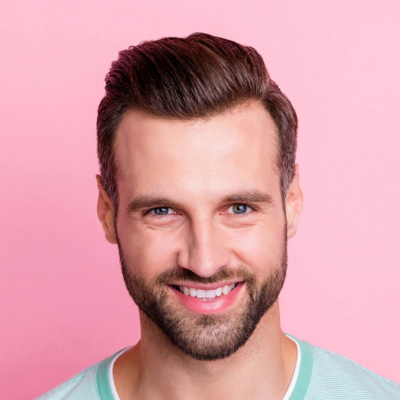 A smiling man with styled hair and a beard against a pink background. He wears a light blue shirt and appears cheerful.