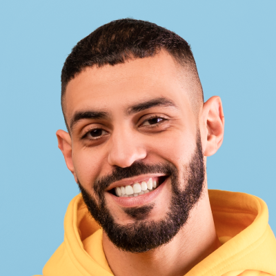 A young man with a beard smiling, wearing a yellow hoodie, against a blue background.