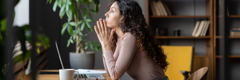 Woman sitting at a desk with a laptop, using the Biteable video maker, looking away thoughtfully in a well-lit room with plants and shelves.