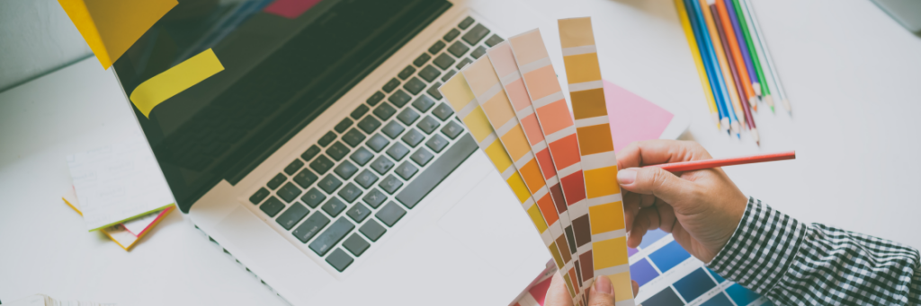 Person holding color swatches with a pencil in hand beside a laptop and colored pencils on a desk.