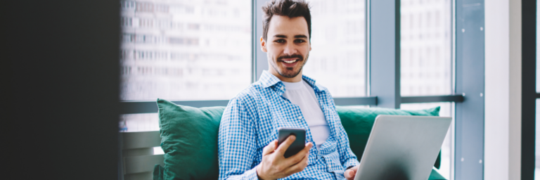 Man in a blue plaid shirt sitting with a laptop on his lap and holding a smartphone, smiling in a modern office with large windows, using the Biteable video maker.