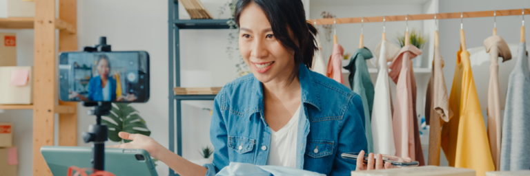 Woman recording a fashion vlog on her smartphone in a studio with clothes in the background.